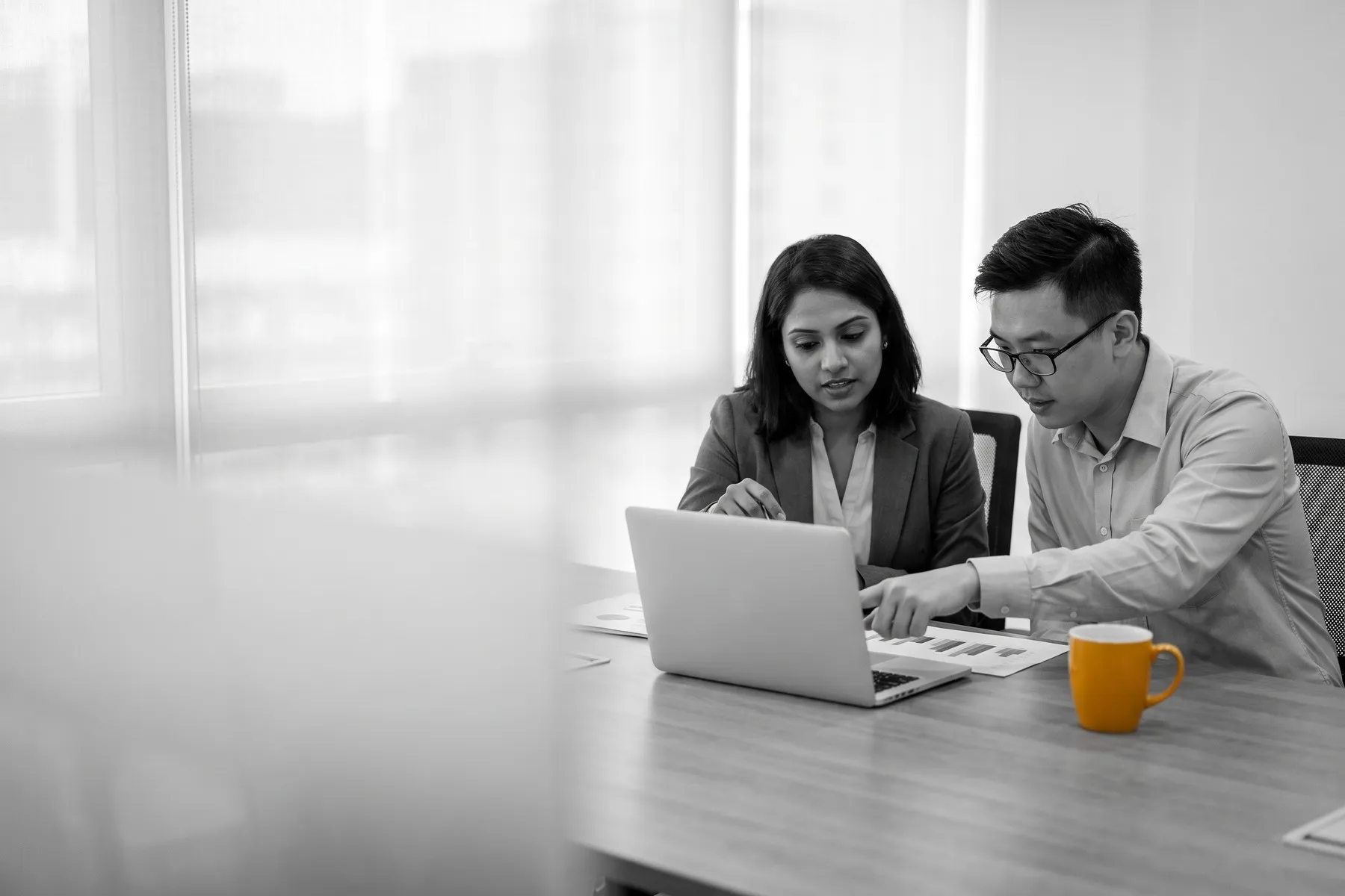 A business owner and a marketing executive review charts on a laptop at a meeting table, beside a yellow coffee mug