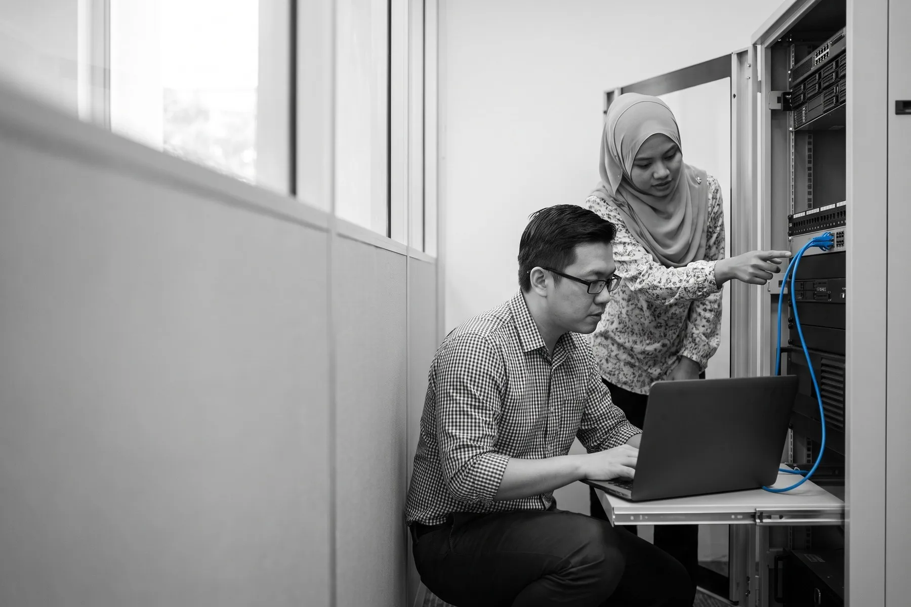 A system administrator checks a laptop beside an open server rack while a colleague points to a blue network cable